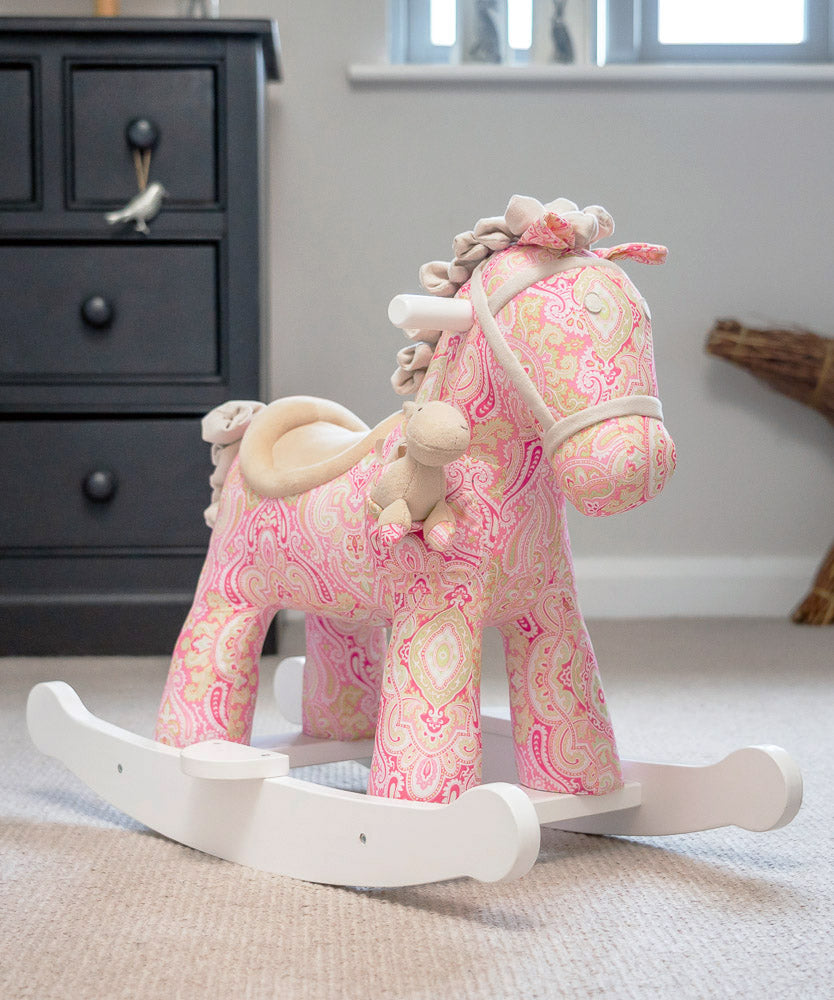 A pink and cream patterned fabric rocking horse toy with a white wooden base, styled in a child's room setting.