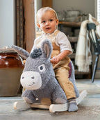 A toddler sits on a plush gray donkey rocking horse in a rustic indoor room with wooden furniture and a tiled floor.