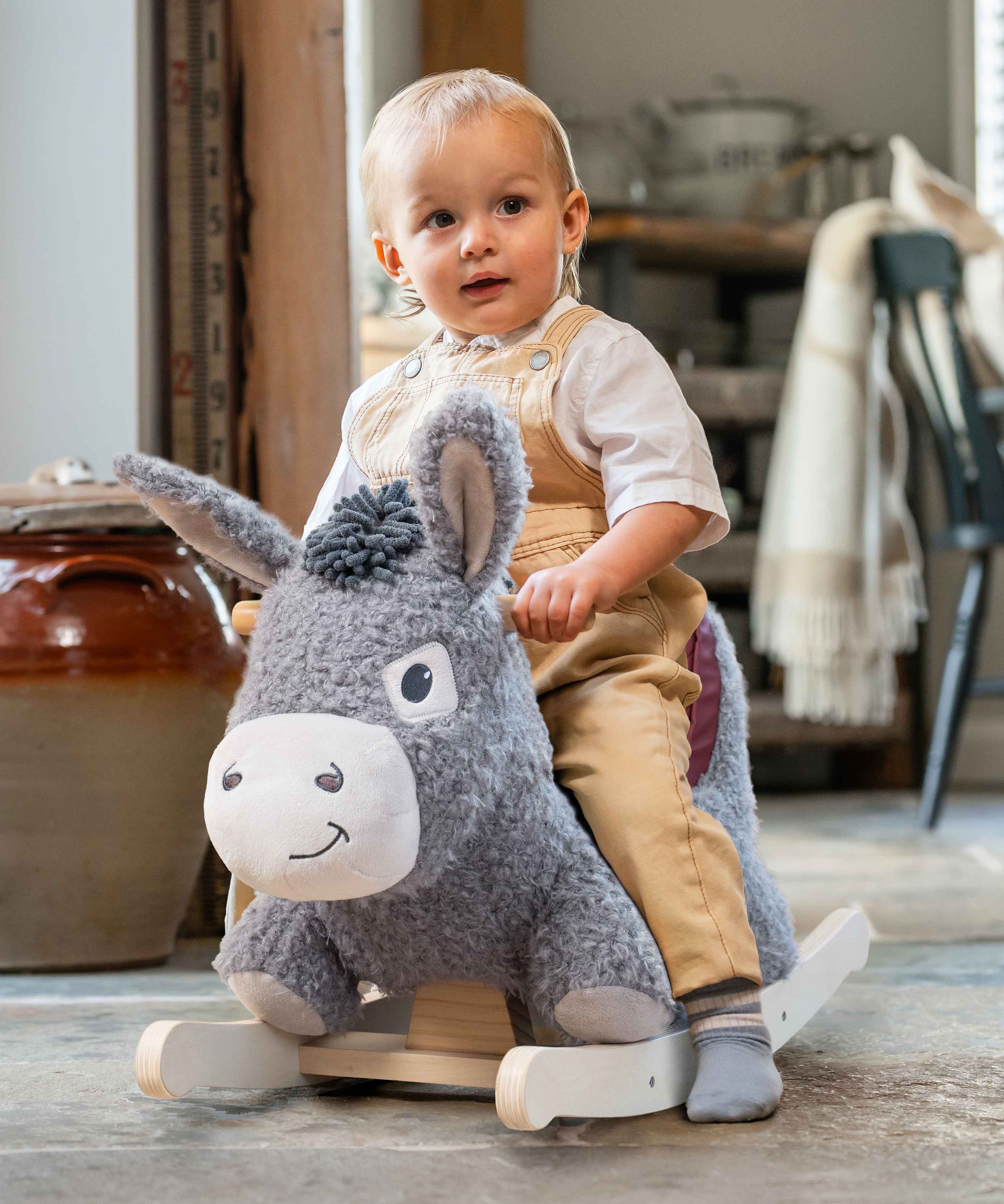 A toddler sits on a plush gray donkey rocking horse in a rustic indoor room with wooden furniture and a tiled floor.