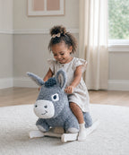 A young child with curly hair wearing a light beige dress sits on a plush gray donkey rocking horse toy in a bright, modern room with beige walls and a white rug.