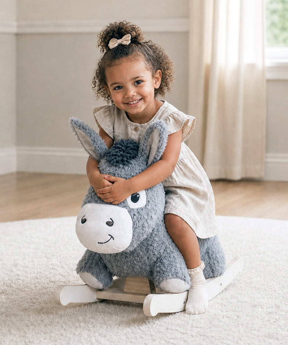 A young child with curly hair wearing a cream dress sits happily on a plush gray rocking donkey toy in a bright, neutral-toned room.