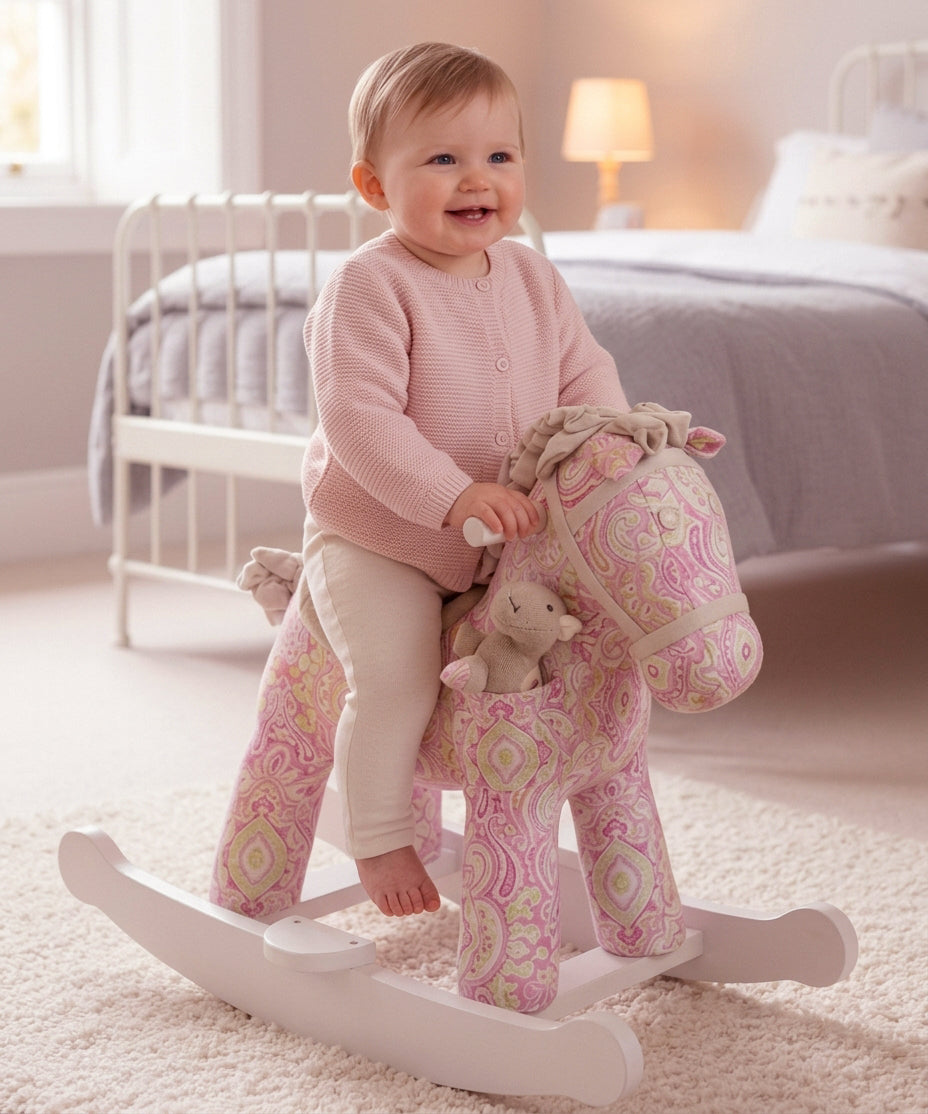 A baby wearing a pink cardigan and cream pants sits smiling on a pink paisley rocking horse toy in a softly lit bedroom.