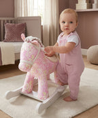A baby in pink overalls stands beside a pink patterned rocking horse toy in a softly lit nursery.