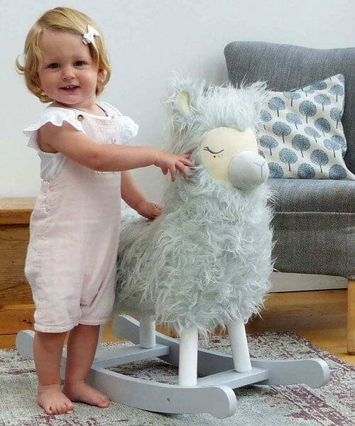 Little girl stands with her Rio Rocking Llama toy in front of a chair