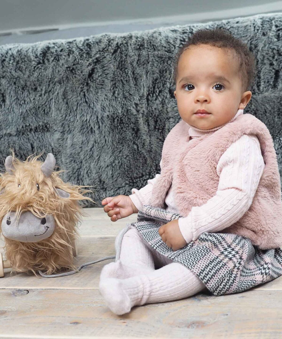 Baby girl sat next to Hubert Highland Cow Pull Along Toy with removable wooden wheels