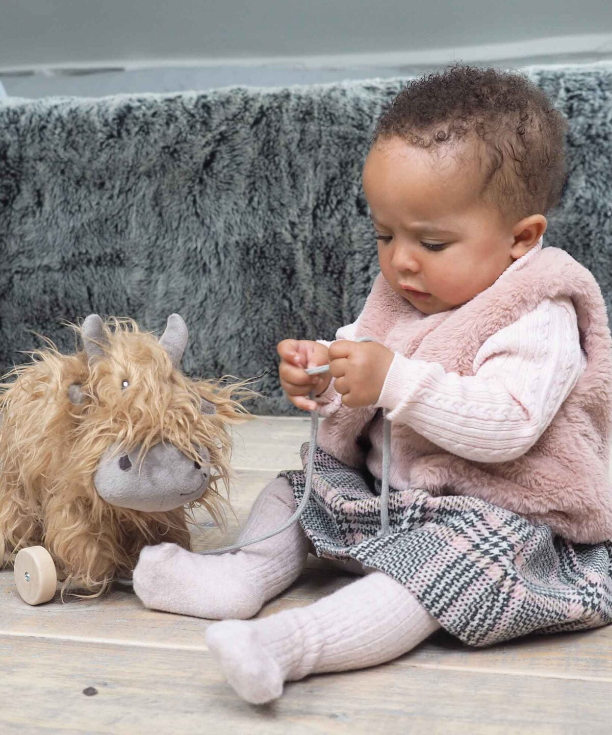 Little girl sits playing with Hubert Highland Cow Pull Along Toy with a thick flowing coat