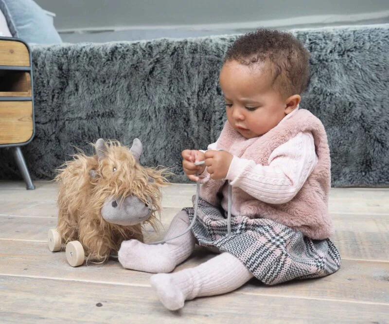 Little girl sits playing with Hubert Highland Cow Pull Along Toy with a thick flowing coat