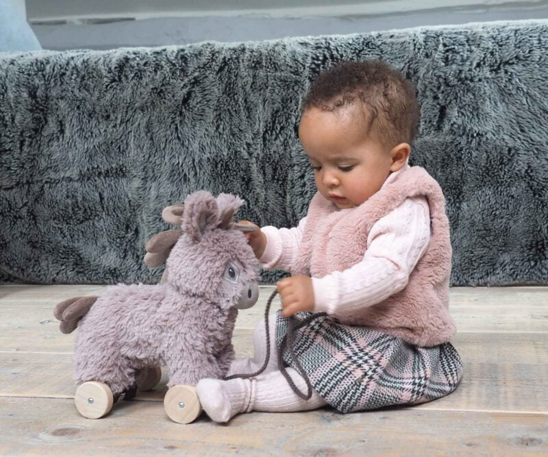 Infant girl sits on the floor playing with Norbert Donkey Pull Along Toy with removable wheels