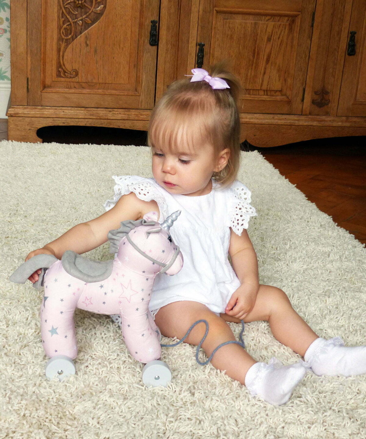 Baby Girl sat on white rug with Celeste Unicorn Pull Along Toy with pink star printed fabric