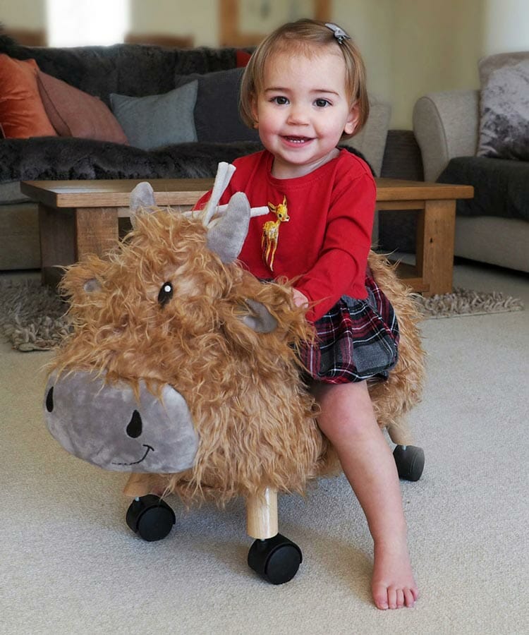 Little girl riding Hubert Highland Cow Ride On Toy with flowing coat Wheeled toys for babies